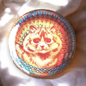Op de afbeelding: Een ronde keramische onderzetter met een kleurrijk mandala-ontwerp met een kat in het midden. De kat is oranje en wit met een gedetailleerde illustratie van zijn vacht. Het mandala-ontwerp is in tinten rood, oranje, geel, blauw en groen.