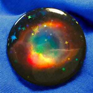 Può includere: Un bottone rotondo nero con un'immagine olografica di una nebulosa con stelle e un centro dai colori dell'arcobaleno.