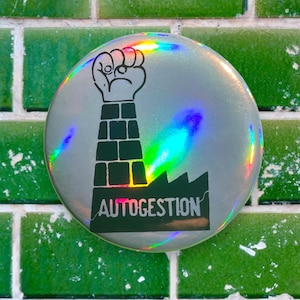 Puede incluir: Un botón holográfico plateado con una ilustración en blanco y negro de un puño levantado rompiendo una pared de ladrillos. El texto "AUTOGESTION" está impreso debajo de la ilustración.
