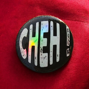 Könnte beinhalten: Ein schwarz-weißer Knopf mit dem Wort "CHEH!" in weißen Buchstaben mit einem regenbogenfarbenen, schillernden Effekt.