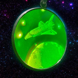 Peut inclure: Un porte-clés en verre iridescent vert avec une navette spatiale en orbite autour d'une planète. Le porte-clés est rond et possède un cadre en métal argenté.