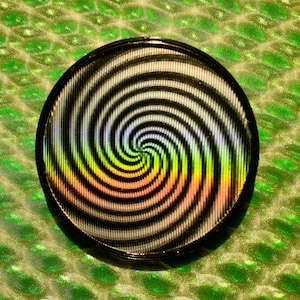 May include: A circular object with a black border featuring a hypnotic spiral pattern. The spiral transitions from black and white to a spectrum of colours, including yellow, orange, and pink. It sits on a textured green background.
