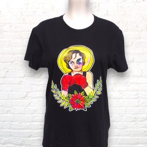 Könnte beinhalten: Schwarzes T-Shirt mit einer bunten Illustration einer Boxerin, die rote Boxhandschuhe trägt. Die Illustration ist von grünen Blättern und einer roten Blume umgeben.
