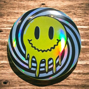 May include: A yellow smiley face with a dripping effect, set against a black and white swirling background. The smiley face is on a round, iridescent button.