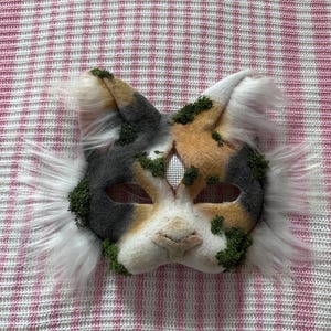 May include: A handmade cat mask with a calico pattern in gray, tan, and white. The mask features faux fur accents and is adorned with green moss. The mask is designed to be worn and has eye holes for visibility.