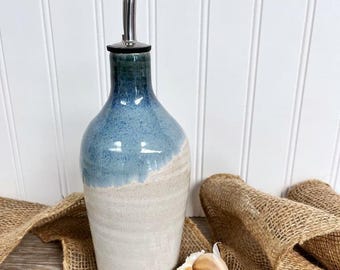 May include: A ceramic oil cruet with a tapered neck and a stainless steel pour spout. The bottle features a two-tone glaze with a deep blue top section that transitions into a textured, off-white base. Three garlic cloves sit in front.