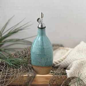 May include: A turquoise ceramic oil bottle with a stainless steel spout. The bottle has a speckled glaze and a natural, unglazed base. The background includes greenery, a wooden crate, and a fishing net.