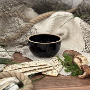 May include: A black ceramic bowl with a light brown rim, set on a wooden surface. The bowl is surrounded by crackers, mushrooms, and greenery. A rope and netting are in the background, suggesting a rustic theme.
