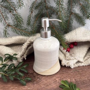 May include: A ceramic soap dispenser with a stainless steel pump. The dispenser is primarily off-white with a tan accent on the lower portion. The background includes greenery and a folded, textured cloth.
