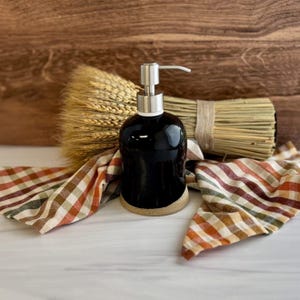 May include: A black ceramic soap dispenser with a silver pump, set on a beige base. The dispenser is surrounded by a bundle of wheat, a bundle of dried grass, and a plaid patterned cloth in shades of orange, brown, and green.