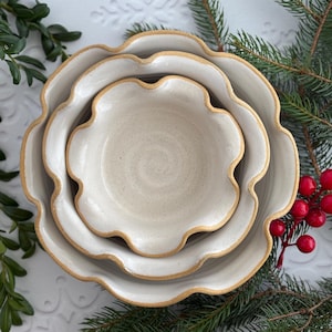 May include: A set of four off-white ceramic bowls with a scalloped edge and a light brown rim. The bowls are nested together, with the smallest bowl in the center. The bowls are surrounded by greenery and red berries.