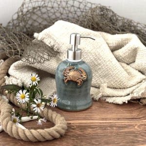 May include: A teal ceramic soap dispenser with a bronze crab design. The dispenser has a silver pump and is surrounded by nautical elements, including rope, flowers, and a fishing net. The dispenser is on a wooden surface.