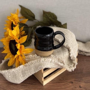 May include: A black ceramic mug with a textured design and a tan base sits on a cream-colored, fringed cloth. Bright yellow sunflowers with green leaves are arranged nearby. The mug has a curved handle and is displayed on a wooden crate.