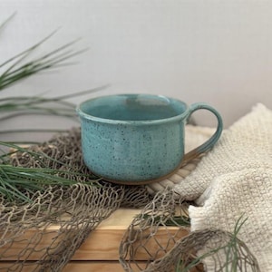 May include: A turquoise ceramic mug with a speckled finish and a rounded handle. The mug sits on a wooden surface with a fishing net and greenery in the background. The mug has a rustic, handmade appearance.