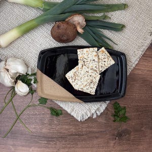 May include: A black rectangular serving dish with a tan corner holds several crackers. Fresh leeks, garlic, mushrooms, and parsley are arranged around the dish on a textured beige surface, creating a rustic food presentation.