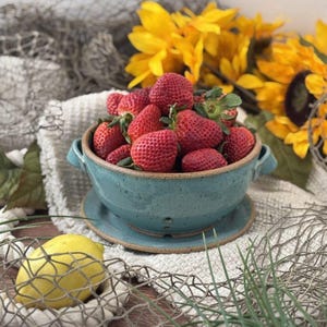 May include: A turquoise ceramic bowl filled with fresh, ripe strawberries. The bowl sits on a matching saucer, with sunflowers in the background. A lemon and decorative netting complete the rustic still life.