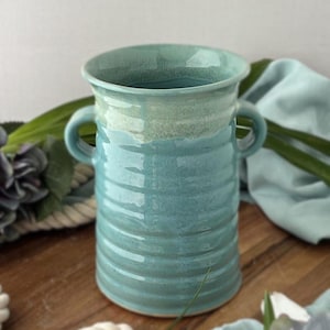May include: A handcrafted, turquoise ceramic vase with two handles. The vase has a wide, flared rim and a textured, ribbed body. The color transitions from a lighter turquoise at the rim to a deeper turquoise at the base. A decorative piece for the home.