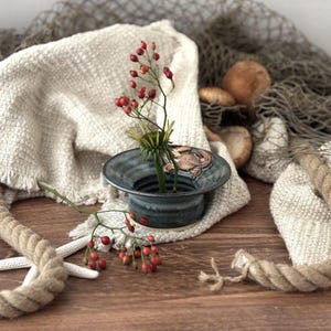 May include: A small, dark blue ceramic vase holds a sprig of red berries and greenery. The vase sits on a textured, off-white cloth, with a starfish and rope nearby. A fishing net and mushrooms are in the background, creating a rustic, nautical theme.