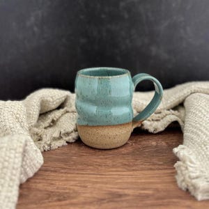 May include: A handmade ceramic mug with a teal glazed upper half and a natural tan base. The mug has a textured, ridged design and a curved handle. The mug is displayed on a wooden surface with a neutral-colored textile in the background.
