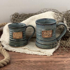 May include: Two ceramic mugs with a blue-grey glaze and horizontal ridges. Each mug features a rectangular tan emblem on the front displaying the Maryland state flag design. The mugs have a sturdy handle and a smooth, glossy finish on the exterior.