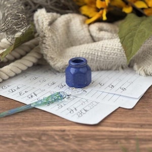 May include: A blue ceramic inkwell sits on lined paper with handwritten calligraphy. A glass dip pen with blue and green accents rests on the paper. The scene includes a neutral-toned cloth and a wooden surface, evoking a vintage aesthetic.
