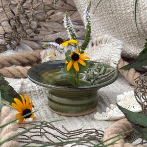 May include: A round, olive green ceramic bowl with a wide, flat rim and a small sculpted turtle figure on the edge. The bowl contains a floral arrangement featuring yellow black-eyed susan flowers and tall, white, spiky botanical stems.
