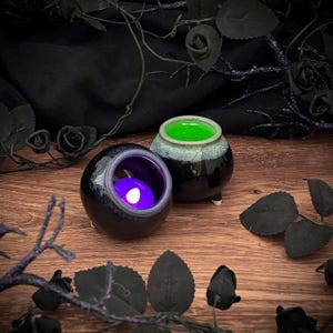 May include: Two small, round ceramic cauldrons with three short legs. One cauldron features a glossy black exterior with a vibrant purple interior containing a glowing light. The other cauldron has a black exterior with a bright green interior.