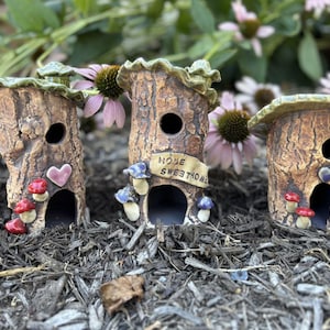May include: Three ceramic fairy houses with a tree trunk design. Each house has a round opening, a heart, and mushroom accents. One house has a banner that reads "HOME SWEET HOME". The houses are brown, red, and blue.