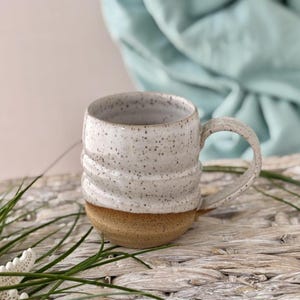 May include: A ceramic mug featuring a speckled white upper section and a solid tan speckled lower section. The mug has a rounded body with two horizontal ridges encircling the middle and a curved handle on the right side for holding.