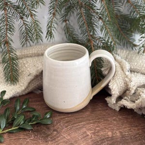 May include: A ceramic mug with a smooth, off-white glazed upper section and a textured, unglazed beige base. The mug features a rounded handle on the right side and subtle horizontal ridges along the interior wall of the cylindrical vessel.