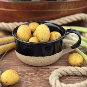 May include: A ceramic mug with a glossy black upper section and a textured, speckled beige base, filled with several small, round, yellow potatoes. The mug features a curved handle on the right side and sits among loose potatoes and rope.
