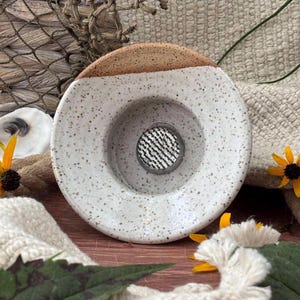 May include: A round, speckled ceramic flower frog with a white glazed basin and a natural tan rim. The center features a circular metal grid insert with a fine mesh pattern, designed to hold flower stems upright in a floral arrangement.