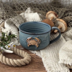 May include: A blue ceramic mug with a crab design. The mug has a handle and a textured surface. The crab is a light brown color and is attached to the front of the mug. The mug is sitting on a white cloth.