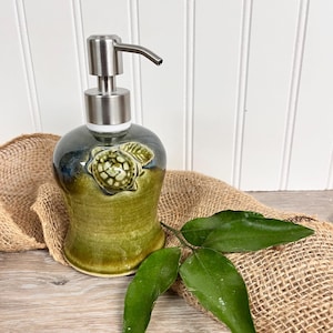 May include: A ceramic soap dispenser with a green and blue glaze. The dispenser features a sculpted turtle design and a stainless steel pump.