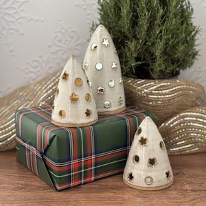 May include: Three ceramic Christmas tree decorations with star and circle cutouts. The trees are off-white with gold and silver accents. They sit on a green and red plaid wrapped gift box. A small potted plant is in the background.