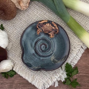 May include: A round, dark blue-grey ceramic dish with an irregular, wavy edge. The center features a raised, embossed brown crab figure and a spiral pattern etched into the glazed surface. The dish has a glossy finish and a dark brown rim.