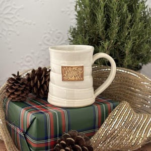 May include: Cream-colored ceramic mug with a handle and a rectangular emblem. The mug is ribbed and sits on a wrapped gift with a green, red, and blue plaid pattern. Pinecones and gold ribbon are also present.