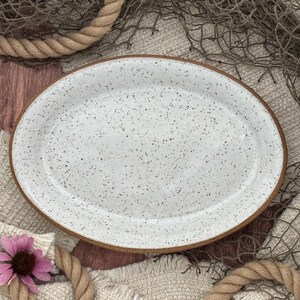 May include: An oval ceramic platter featuring a white glaze with a speckled brown pattern throughout. The platter has a smooth, glossy surface and a distinct, unglazed brown rim that outlines the entire edge of the dish, creating a contrast.