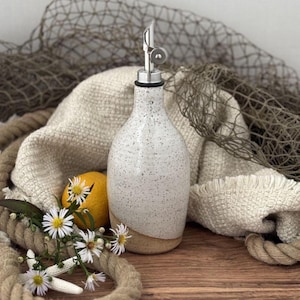 May include: A ceramic oil dispenser with a speckled white glaze on the upper portion and a natural tan base. The bottle features a silver-toned metal pour spout with a hinged cap. A yellow lemon and small white flowers sit nearby.