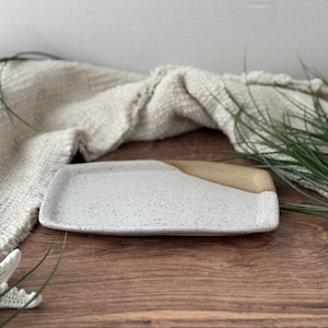 May include: A rectangular ceramic serving tray with a speckled white surface and a tan-colored section on one corner. The tray is set on a wooden surface with a neutral-colored cloth in the background.