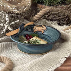 May include: A round, teal-glazed ceramic bowl featuring a sculpted crab on the rim and an integrated wooden spreader. The bowl contains a wedge of cheese garnished with fresh rosemary and cranberries, resting within a coastal-themed display.