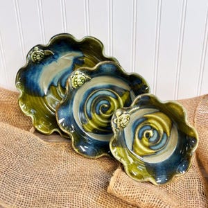 May include: Set of three ceramic bowls with a wavy edge design. The bowls feature a spiral pattern in shades of blue and green, with a small turtle figurine on the rim. The bowls are displayed on a burlap surface.