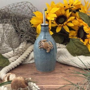 May include: A blue ceramic oil bottle with a stainless steel spout and a bronze crab emblem. The bottle is displayed on a wooden surface with sunflowers and nautical decor in the background.