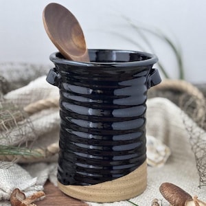 May include: A black ceramic utensil holder with a textured, ribbed design and a natural tan base. A wooden spoon rests inside. The holder has two small handles and a glossy finish, perfect for kitchen organization.