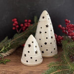 May include: Two off-white ceramic Christmas tree decorations. The trees are cone-shaped with star and dot cutouts. The trees are on a wooden surface with red berries and greenery in the background.