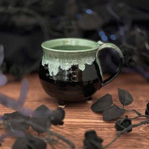 May include: A round ceramic mug with a glossy black base and a textured, light green drip glaze around the rim. The mug features a curved handle on the right side and stands on three small, short feet, creating a cauldron-like silhouette.