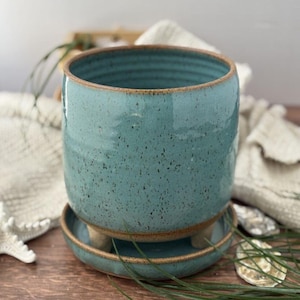 May include: A turquoise ceramic planter with a matching saucer. The planter has a speckled finish and a rounded shape. The saucer is supported by three small feet. The rim of the planter and saucer have a brown trim.