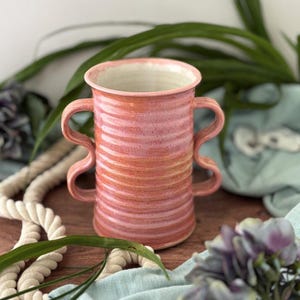 May include: A handmade ceramic vase with a pink and orange gradient. The vase has two unique, curved handles and a textured, ribbed surface. The interior is a light cream color. A decorative piece for home decor.