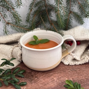 May include: A white ceramic mug filled with orange soup, garnished with a sprig of mint. The mug has a beige handle and sits on a wooden surface, with a beige cloth and greenery in the background.