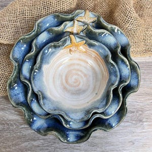 May include: Set of four ceramic nesting bowls with a wavy edge design. The bowls are glazed in shades of blue and white, with a tan interior and starfish accents. The bowls are arranged in a nested configuration.